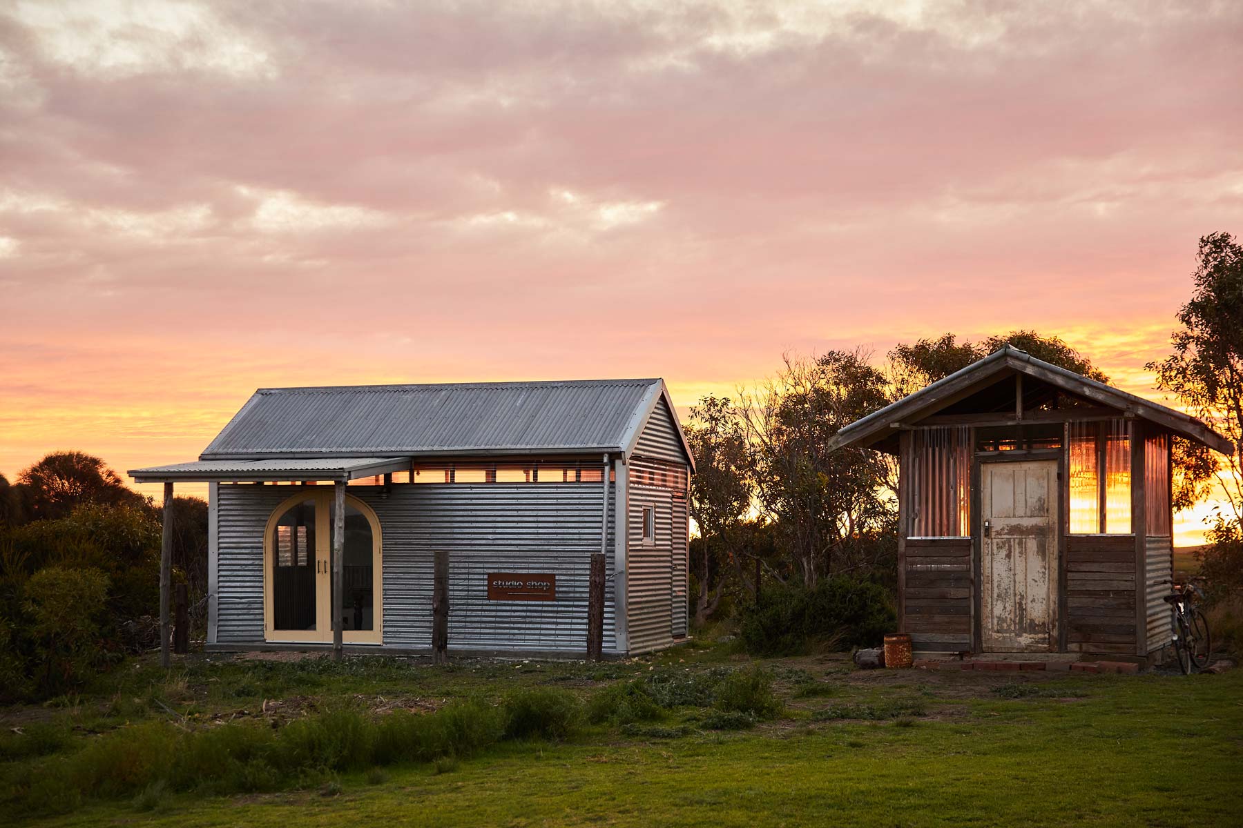 Two rustic wooden cabins at sunrise with a colorful sky. Taken at coorong life on the left is the Art Studio with arch glass doors and the right is the small studio Shop onsite for guest pantry needs, tea coffee etc