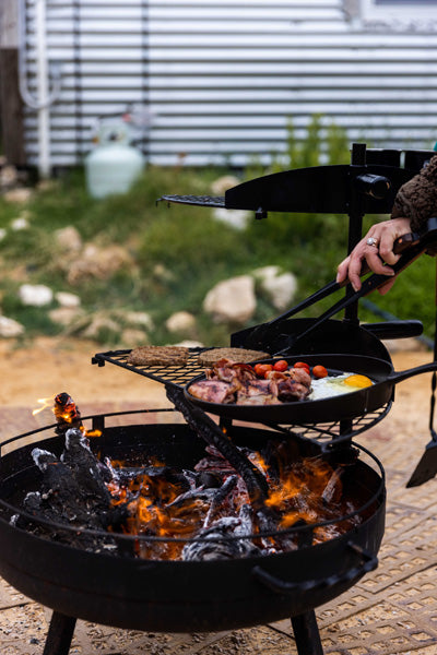 Person grilling food on an outdoor fire grill with a pan over an open flame outdoors. Cooking bacon and eggs and toast and cherry tomatoes