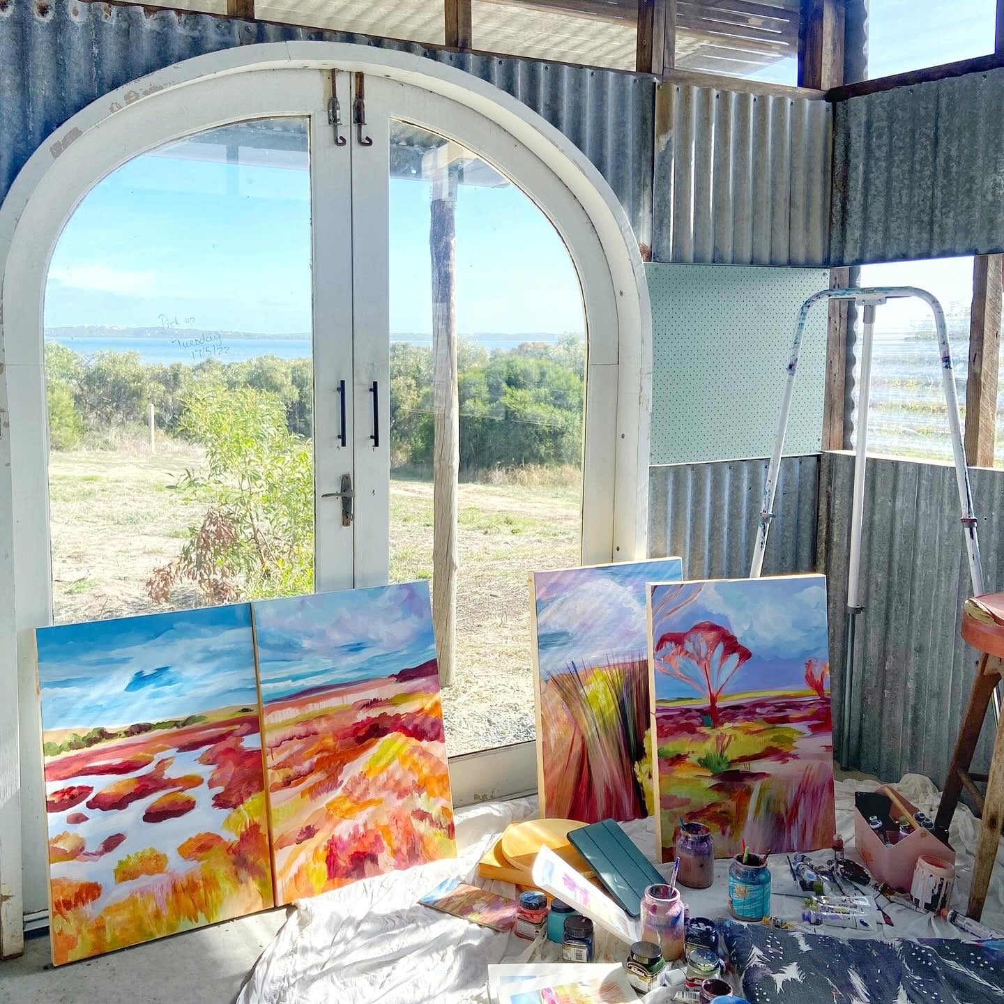 Art studio with paintings and art supplies near a large window with a view of greenery. Art Residency program at Coorong.life