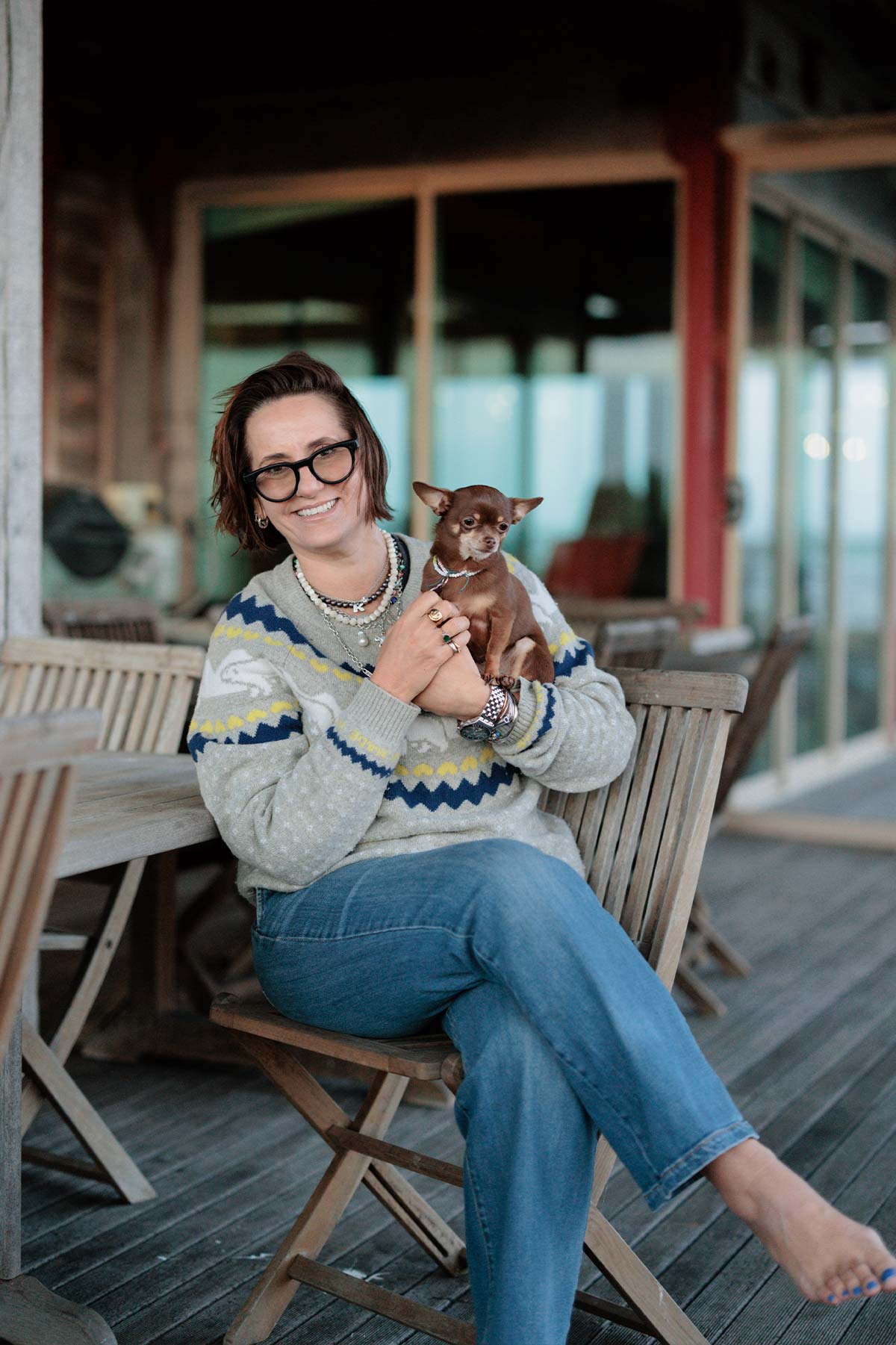Woman sitting on a wooden chair holding a small dog on a deck.