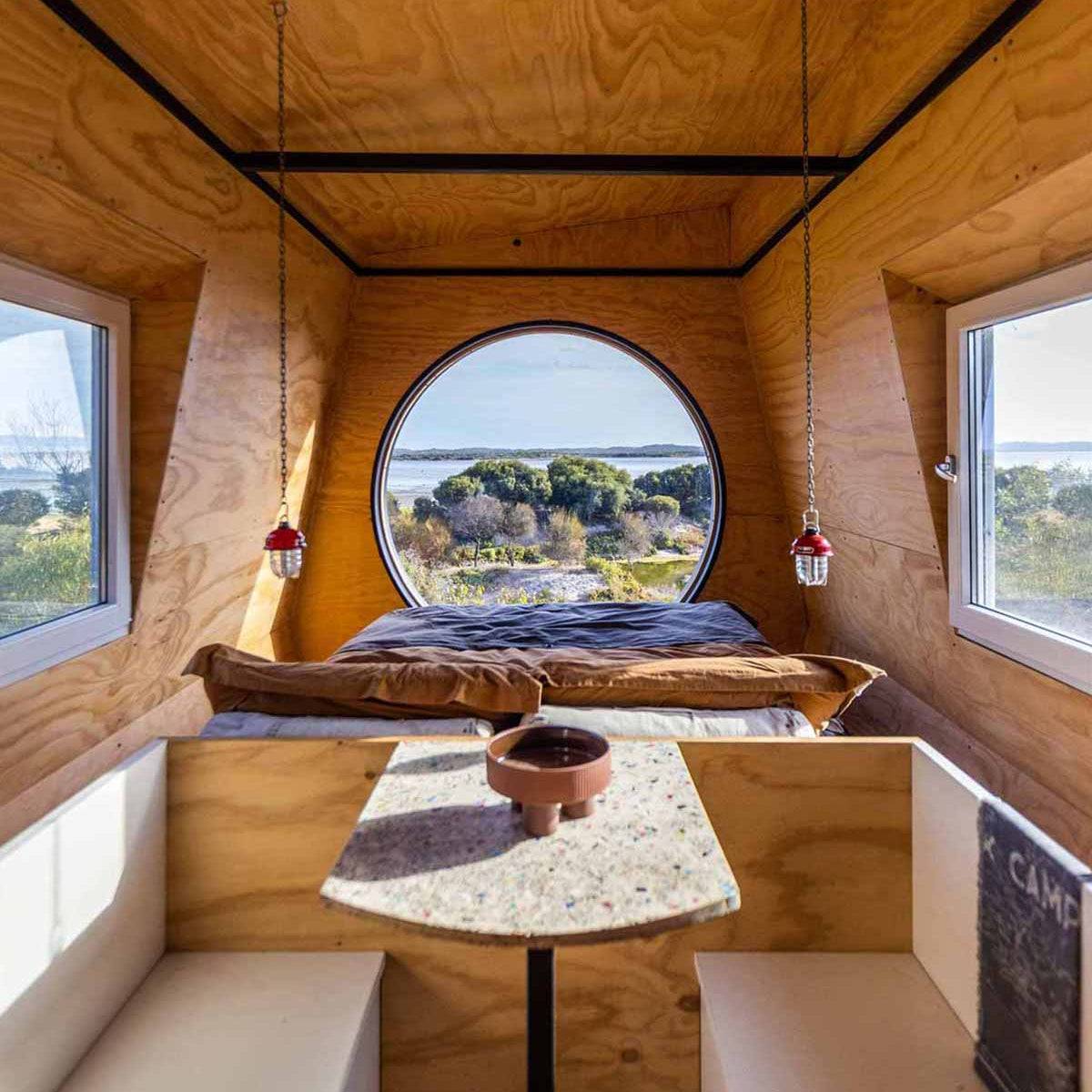 Wooden interior of a compact living space with a round window, bed, and small table. Inside KipPod II Coorong Life