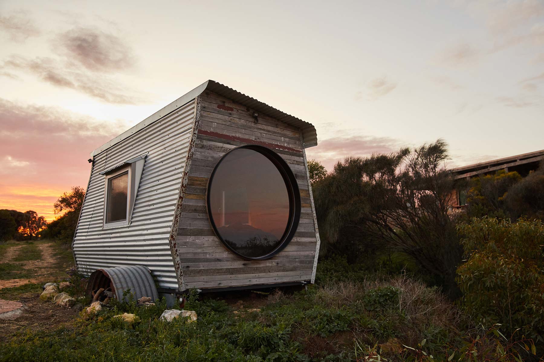 Small wooden cabin at Coorong life called KipPod II with a round window in a natural setting during sunrise. Lovely colours and reflection on window, The Coorong, South Australia
