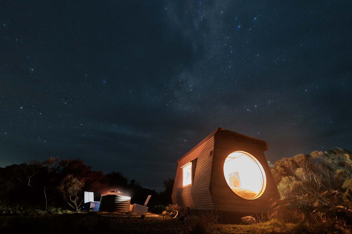 Wooden cabin is one of Coorong Life’s KipPods with night skywith glowing windows under a starry night sky you can see the Milky Way and the lights inside