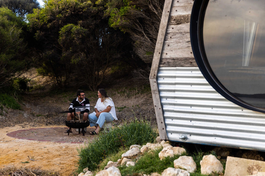 Two people sitting on a bench near KipPod I building with a corrugated metal exterior next to fire pit chatting