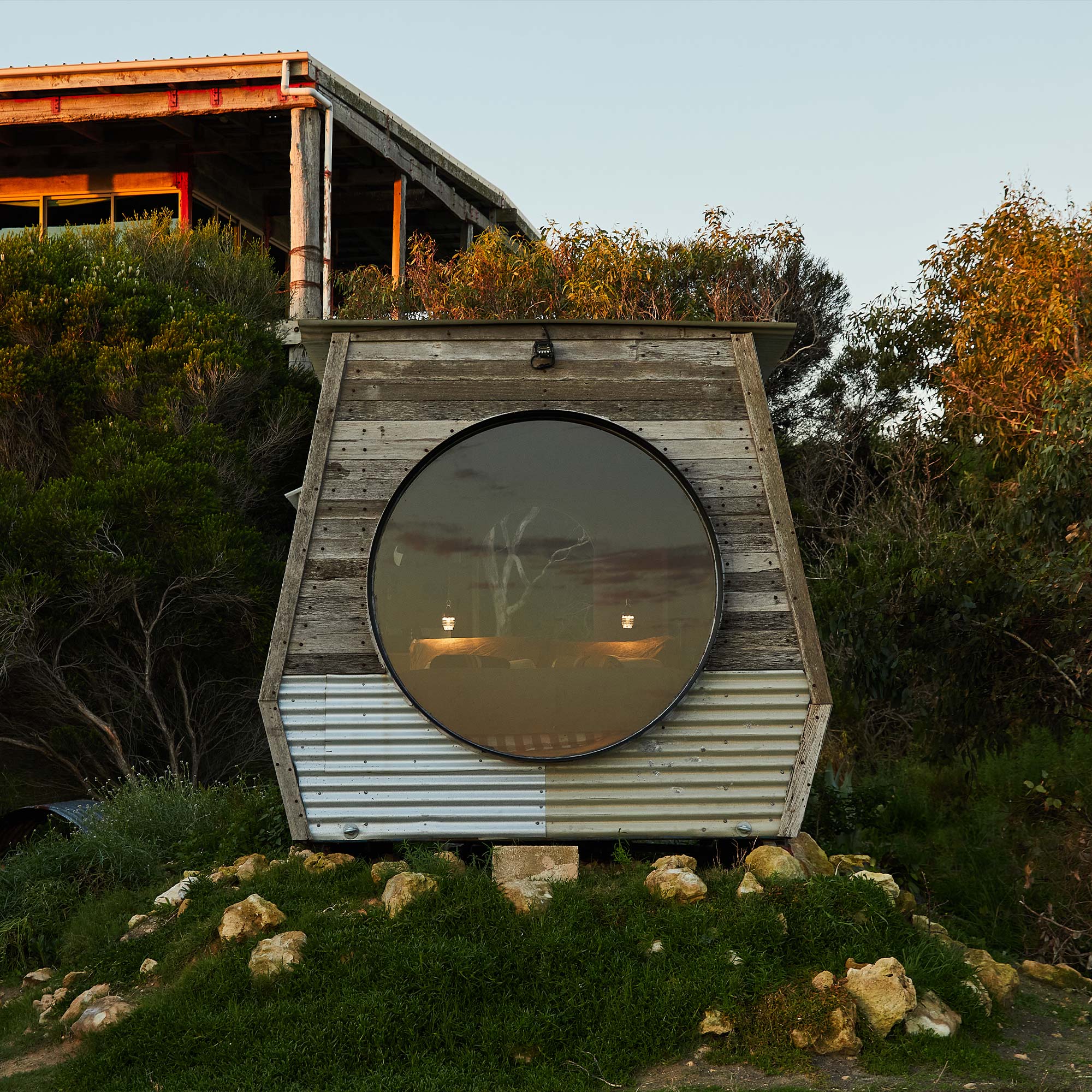 This is a photo of the rustic custom made Pod for accomadation at Coorong life its taken an sunset with the large circle window showing the queen bed and the two night light on, KipPod is surrounded by greenery 