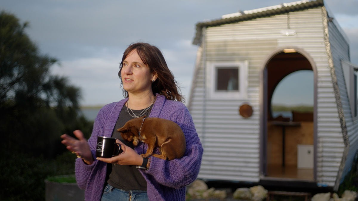 Woman in photo is Kate Brooks owner of Coorong life holding a dog Brandi Lee her co-host and a mug in front of a small house she calls a KipPod 